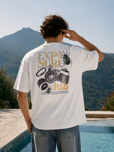 A man viewed from behind wearing a white graphic t-shirt with a camera-themed print and blue jeans, standing next to a swimming pool with mountains in the background.