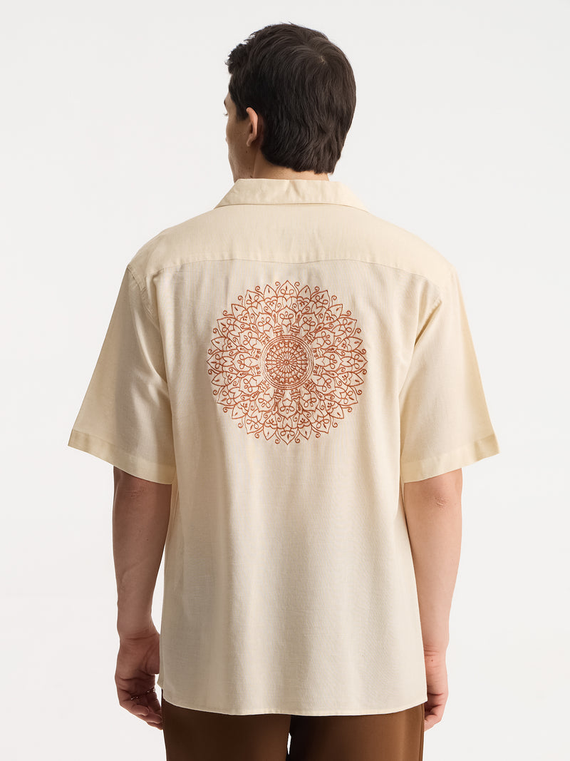 A man viewed from the back wearing a cream-colored short-sleeve button-up shirt with a large circular mandala pattern printed in rust orange on the back, paired with brown pants against a plain white studio background.