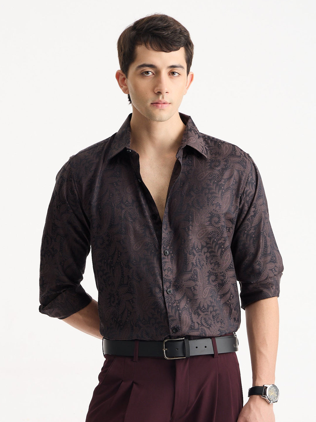 A man wearing a dark brown patterned button-down shirt with rolled-up sleeves, paired with burgundy trousers and a black belt, standing against a plain white background.
