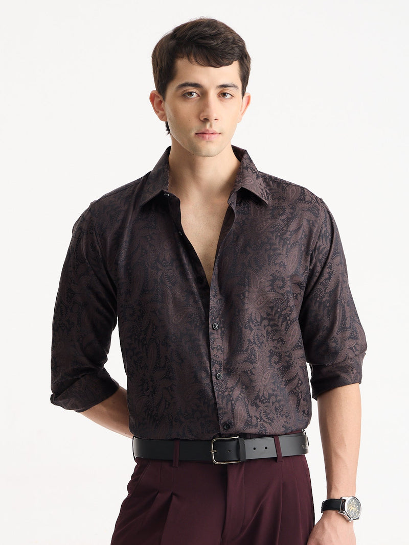 A man wearing a dark brown patterned button-down shirt with rolled-up sleeves, paired with burgundy trousers and a black belt, standing against a plain white background.
