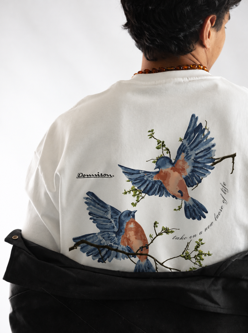 A person viewed from behind wearing a white oversized T-shirt with a large back graphic of two blue birds perched on branches, paired with a black jacket draped over the shoulders against a light gray studio background.
