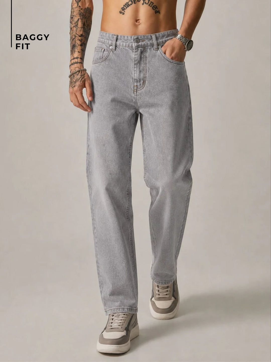 A model wearing light gray baggy-fit jeans, shown from the waist down against a neutral studio background. The model has tattoos on one arm, a watch on the wrist, and is wearing gray and white sneakers.