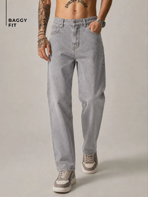 A model wearing light gray baggy-fit jeans, shown from the waist down against a neutral studio background. The model has tattoos on one arm, a watch on the wrist, and is wearing gray and white sneakers.