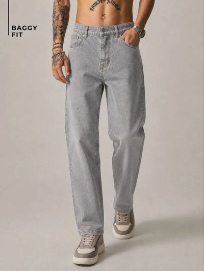 A model wearing light gray baggy-fit jeans, shown from the waist down against a neutral studio background. The model has tattoos on one arm, a watch on the wrist, and is wearing gray and white sneakers.
