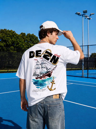 A man on a blue tennis court wearing a white t-shirt with a large nautical graphic on the back, featuring a sailing ship, waves, and an anchor. The shirt has 'DEZN' printed across the upper back in black and gold.