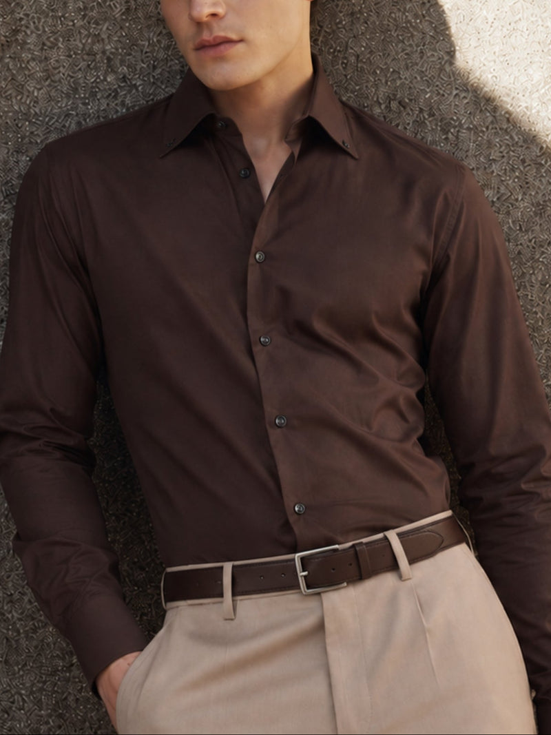 A man wearing a dark brown long-sleeve button-down dress shirt and beige trousers, leaning against a textured concrete wall with dramatic side lighting.