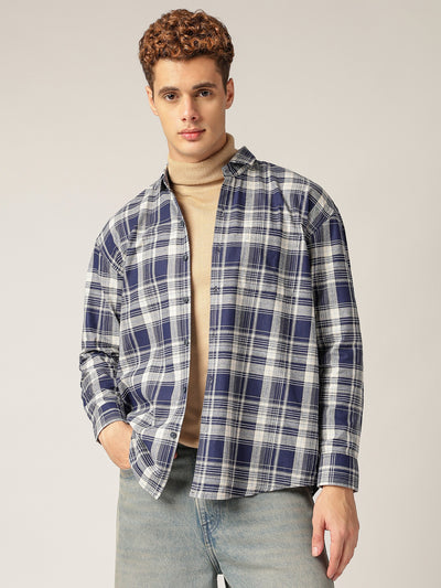 Man wearing a blue plaid shirt over a beige sweater with light blue jeans on a plain background