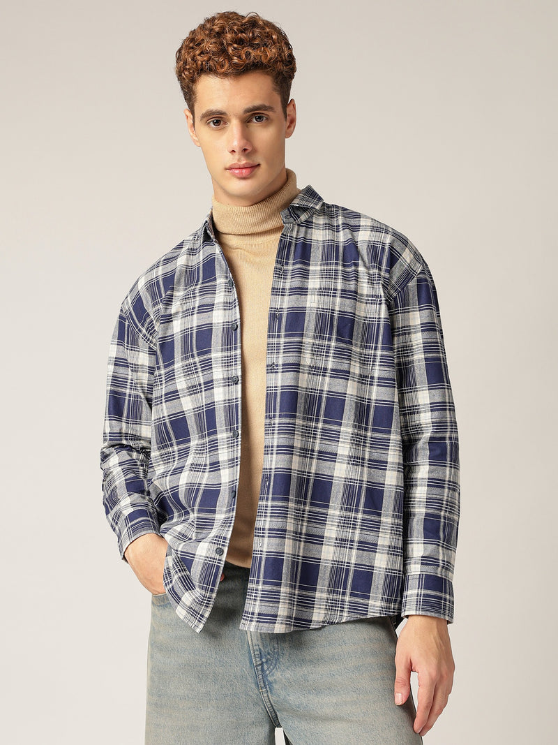 Man wearing a blue plaid shirt over a beige sweater with light blue jeans on a plain background