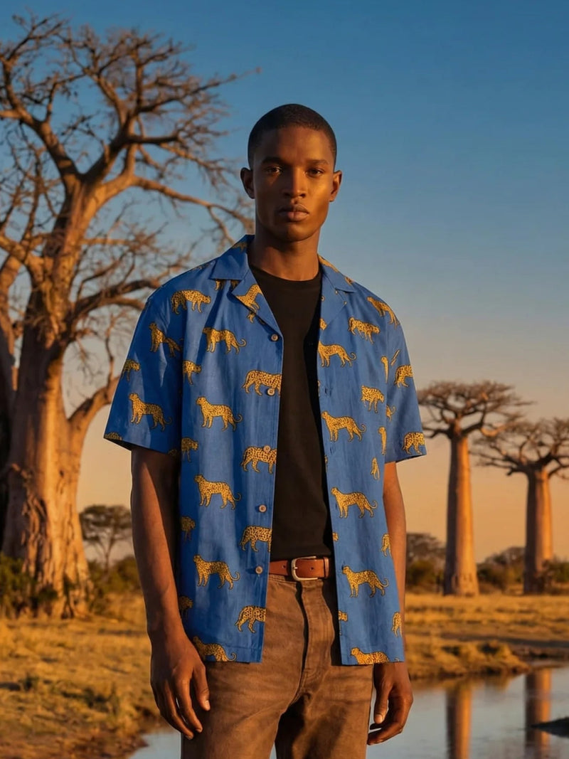 Man wearing a blue shirt with yellow patterns standing in a natural landscape with trees and water.
