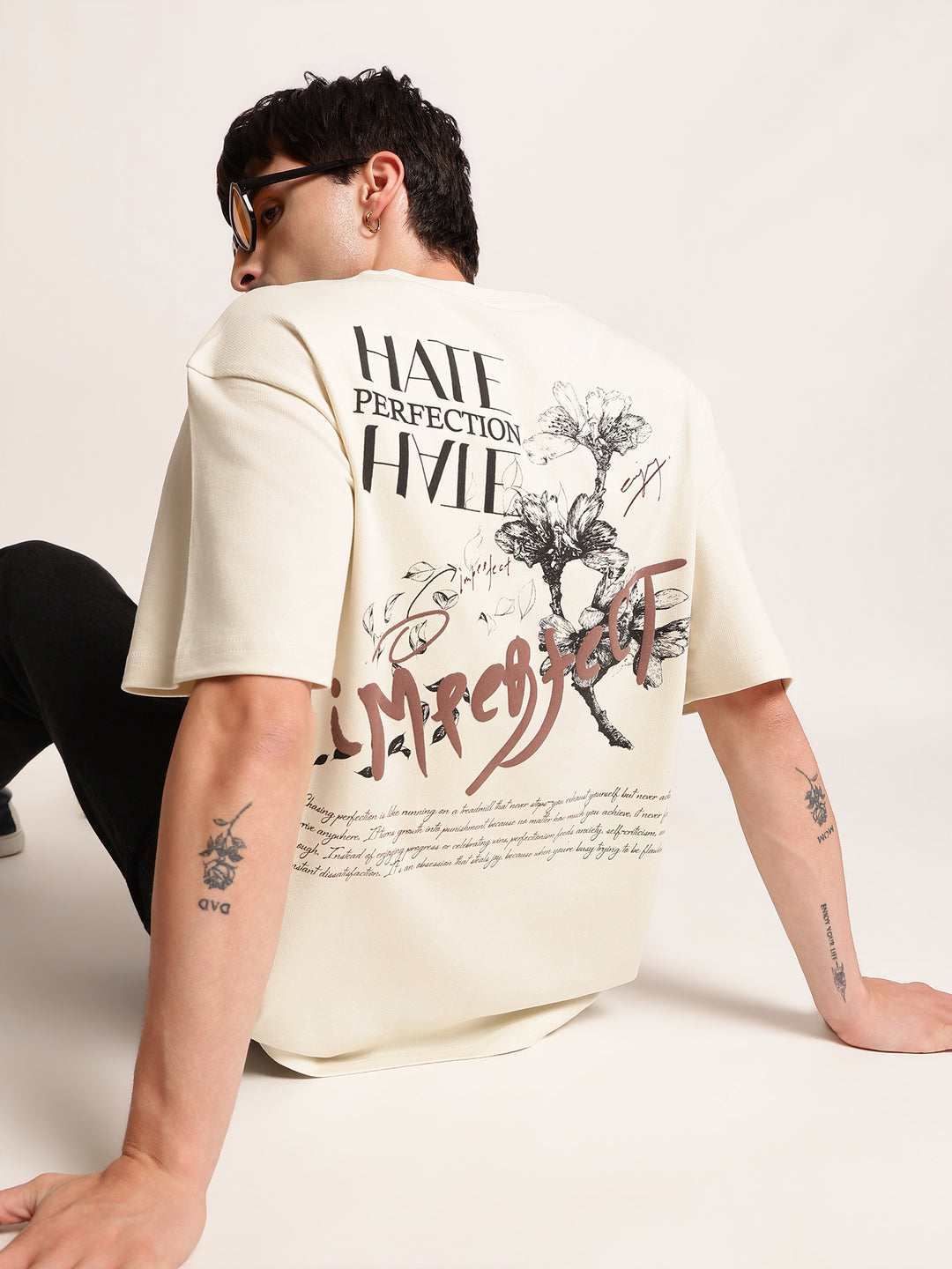 A man wearing a cream-colored graphic t-shirt, shown from behind. The shirt features black text reading 'HATE PERFECTION HATE' and 'Mr.