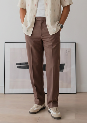 Man wearing a light shirt, brown pants, and sunglasses indoors with a white wall and framed picture in the background.